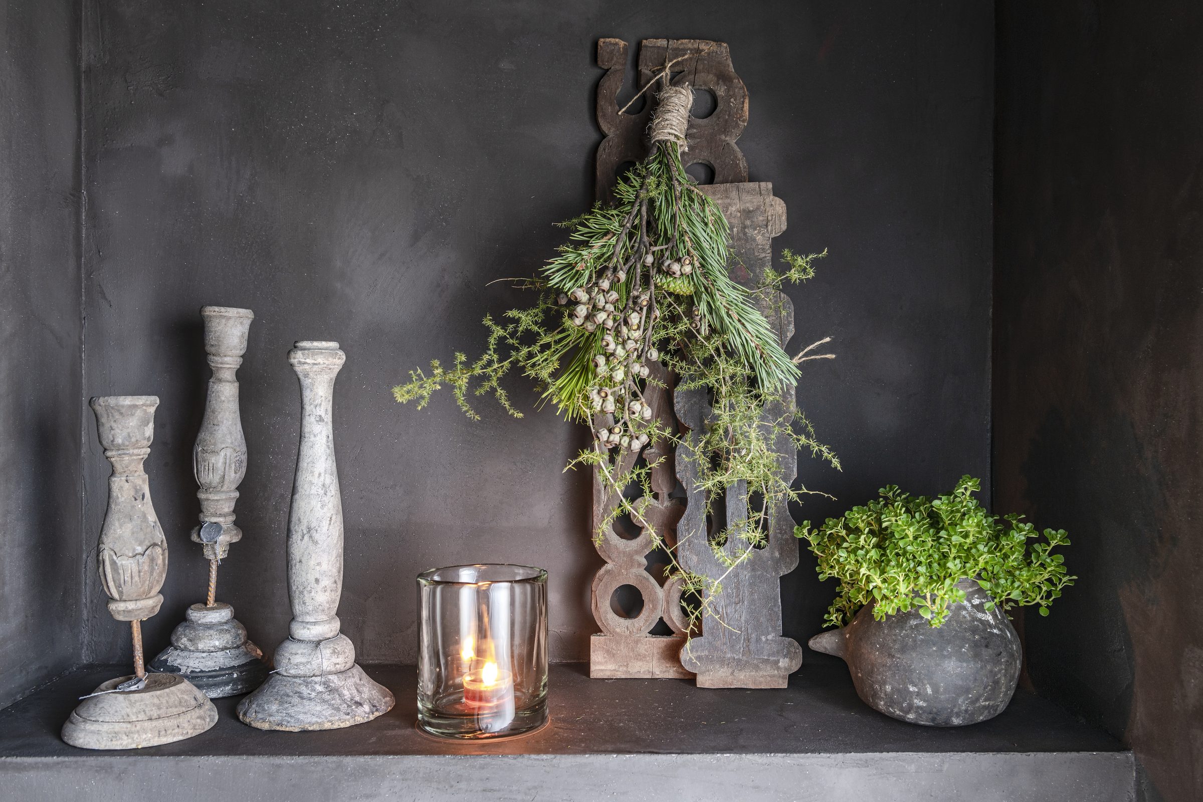 Stilleven met stenen kandelaar, windlicht met brandende kaars en oud houtsnijwerk met groene toef tegen een donkere wand