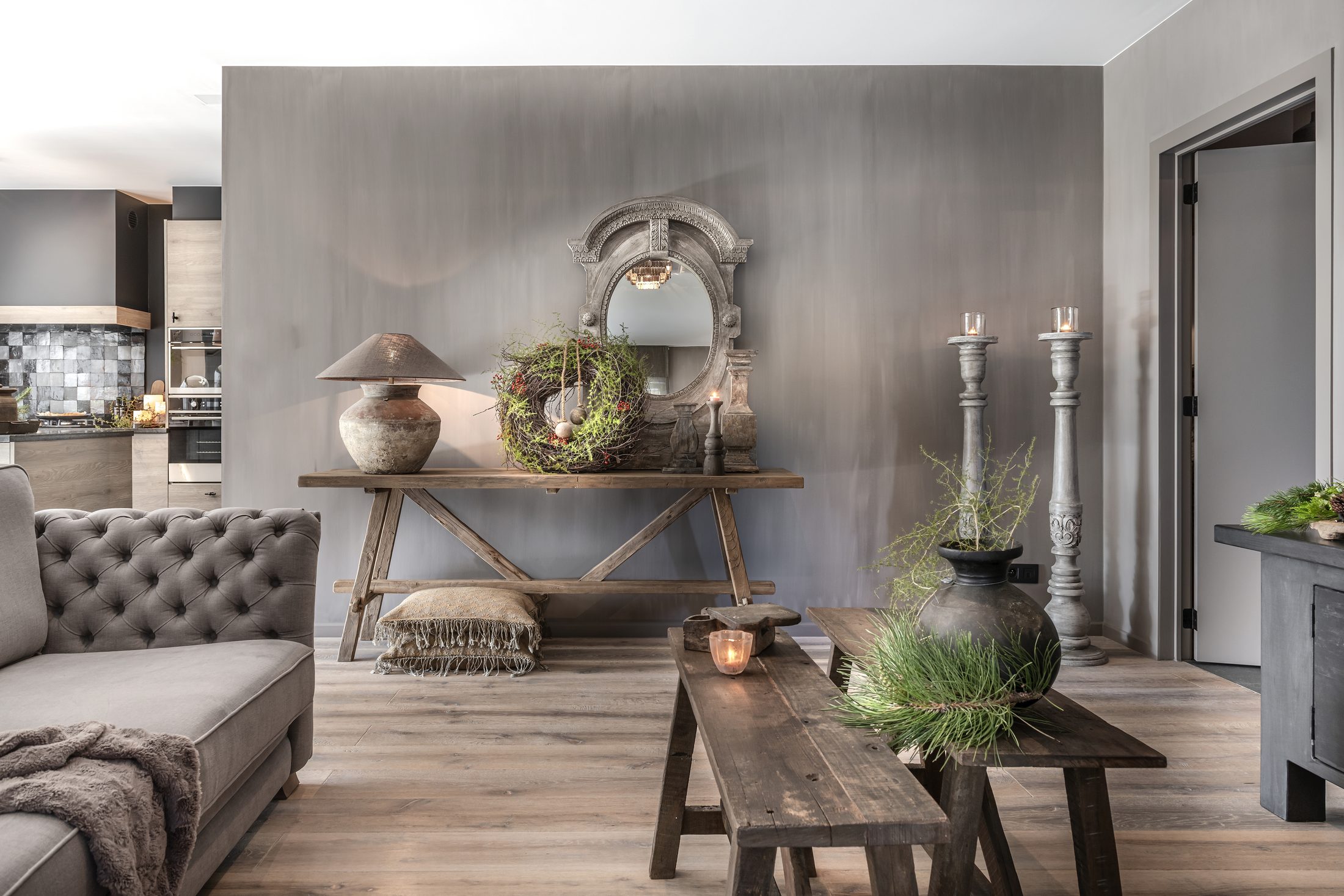 Wandtafel in oud hout met stenen lamp, sierkrans en spiegel, grote kandelaars, een houten bank en een kruik met groen